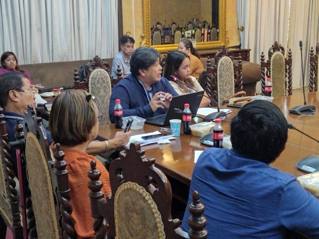 People seated around a wooden conference table with laptops and documents, engaged in a discussion.