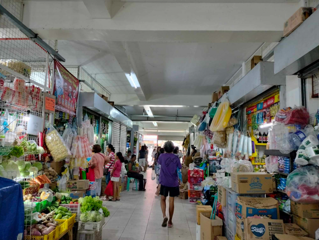 Busy market aisle with vendors selling produce and goods, shoppers browsing, and bright overhead lighting.