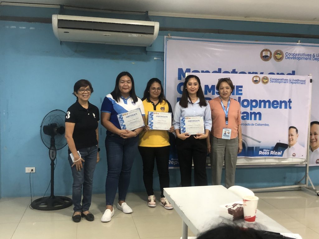 Four women stand together in a room, smiling and holding certificates. They are positioned in front of a banner that promotes a "Cooperative and Livelihood Development Program." The woman on the far left wears a black shirt with the number 3 on it, while the others wear shirts of various colors. The setting appears to be an official event.