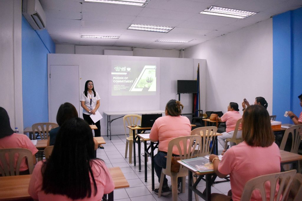 A presenter stands in front of a screen, while an audience in pink shirts engages in a classroom setting.