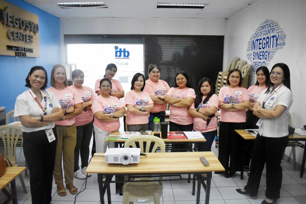 A group of twelve women in matching pink shirts pose with crossed arms in a training center.