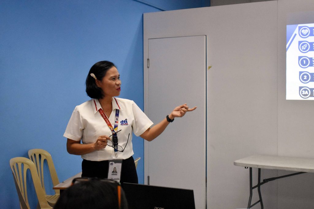 A woman gives a presentation, gesturing towards a screen while speaking to an audience