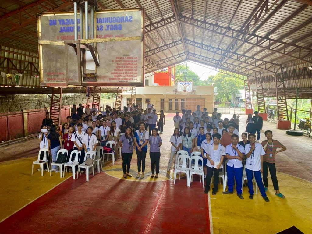 A group of senior high students from San Cristobal Integrated High School attending an entrepreneurship seminar conducted by the Negosyo Center San Pablo City. The seminar is held in a covered gymnasium with students seated and standing, listening attentively. The event aims to equip students with the skills needed to consider entrepreneurship as a career path.