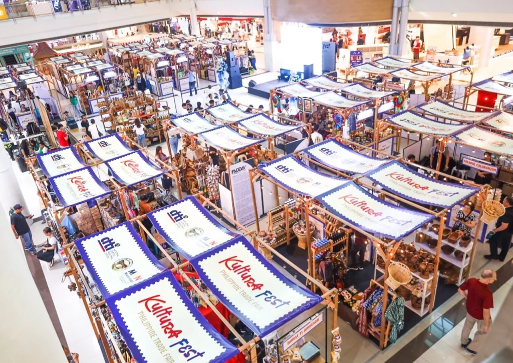 An aerial view of Kultura Fest: Philippine Trade Fair at Festival Mall, Alabang, featuring wooden booths adorned with banners from the DTI and Muntinlupa City Government. Various local products, including handicrafts, food, and fashion items, are displayed as shoppers explore the vibrant marketplace.