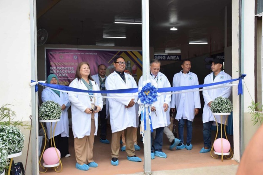 A ribbon-cutting ceremony marks the official turnover of the Shared Service Facility (SSF) for Packaging and Labeling at MSU-Maigo, Lanao del Norte. Officials from DTI Lanao del Norte, MSU-Maigo, and the Provincial Government, dressed in white lab coats and blue shoe covers, stand in front of the facility entrance with a blue ribbon and decorative elements.