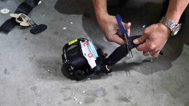 A person is cutting a safety belt with a pair of scissors. The safety belt is part of a batch of substandard safety belts being destroyed under the supervision of the Department of Trade and Industry (DTI) in Taguig City, Philippines. This action is part of a measure to halt the distribution of non-compliant safety belts and ensure consumer protection. The destruction process includes cutting and hammering over 70 pieces of three-point safety belts that failed to meet the corrosion test requirements under Philippine National Standards (PNS) 1892:2000 Amd. 01:2002.