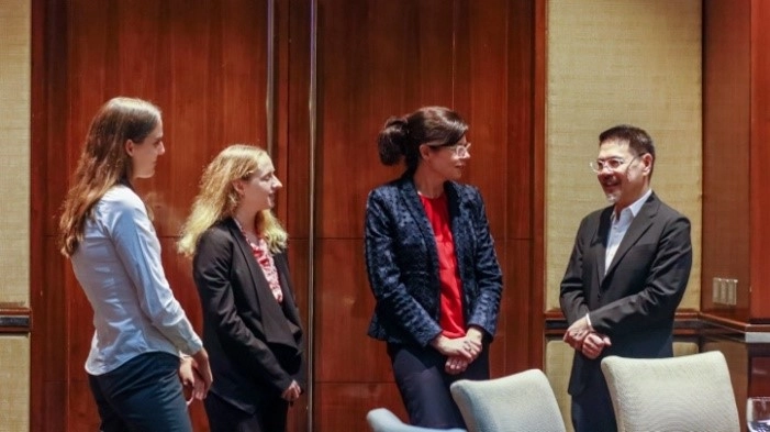 Four individuals are standing in a room with wooden panel walls, engaged in conversation. The individuals are dressed in business attire, suggesting a formal or professional setting.