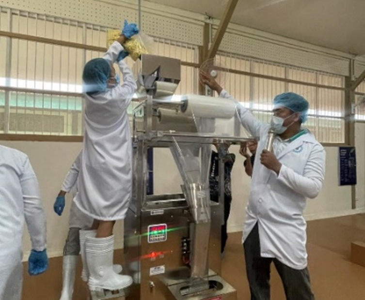 A staff member in a white lab coat, gloves, and boots loads food products into an automated packaging machine while another staff member explains the process using a microphone. Both are wearing hairnets and masks inside the Shared Service Facility (SSF) for Packaging and Labeling at MSU-Maigo, Lanao del Norte.