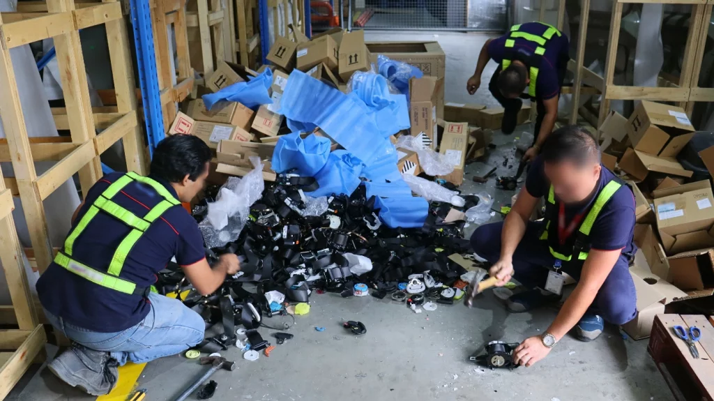 Three individuals wearing high-visibility vests are seen in a warehouse, actively destroying safety belts. The floor is scattered with various parts of the belts, and numerous cardboard boxes are visible in the background. This activity is supervised by the Department of Trade and Industry (DTI) to ensure that non-compliant safety belts, which failed to meet the corrosion test requirements under Philippine National Standards (PNS) 1892:2000 Amd. 01:2002, are removed from the market to protect consumer safety.