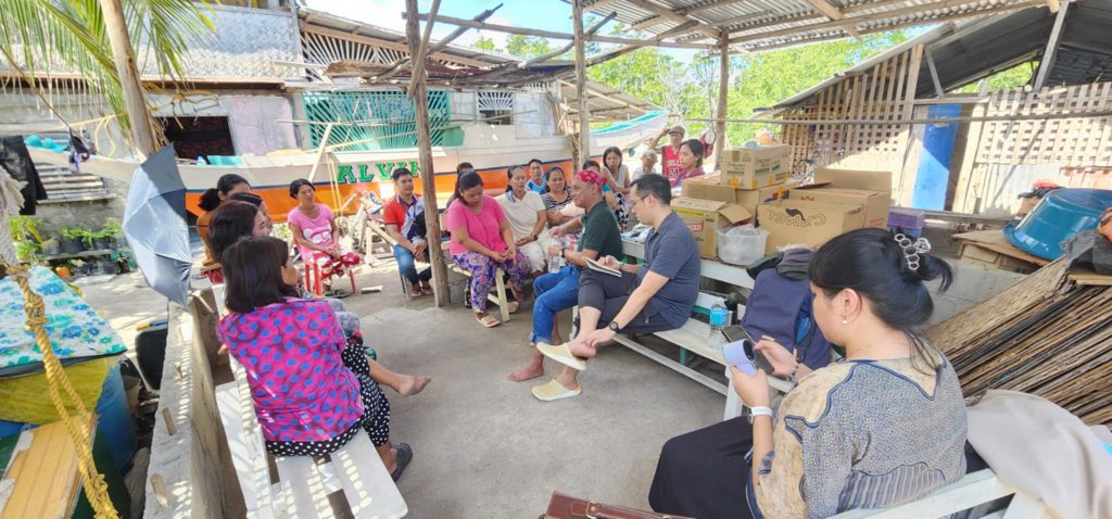 A group of people seated in a semi-circle under a makeshift shelter, participating in a focus group discussion. The setting appears to be a coastal area with a boat visible in the background. Various boxes and supplies are stacked on a table, and some participants are taking notes or holding papers. The focus group, conducted by the Department of Trade and Industry and the Asian Development Bank, took place from 6-7 February 2025 in Roxas, Palawan, and is focused on potential investment projects for the fisheries sector in the municipality.