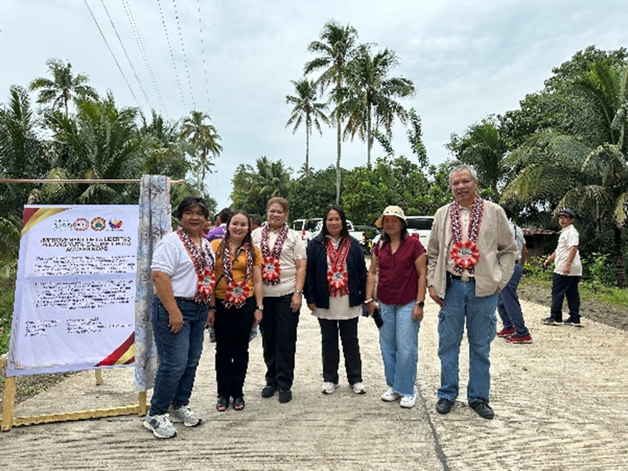 A group of people, including representatives from various local government units and organizations, stand together during the turnover ceremony of a road improvement project. The project reduces travel time and facilitates easier transportation for farmers and local businesses. Key attendees include DTI Region 10 Director Ruel Paclipan and the La Libertad Farmers Multi-Purpose Cooperative (LALIFAMCO).