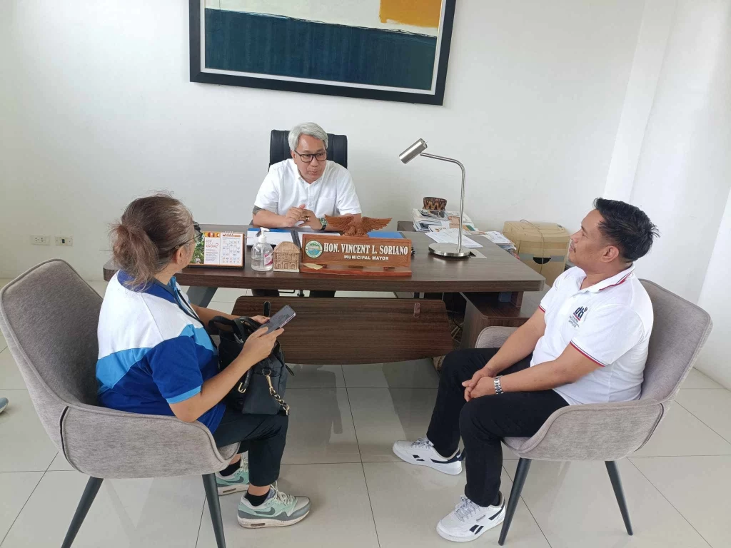 In an office setting, three individuals are seated around a desk. The person behind the desk is Pakil Municipal Mayor Vincent L. Soriano, as indicated by the nameplate on the desk. The two individuals seated in front of the desk are likely officials from the Department of Trade and Industry (DTI). The office is modern, with a framed abstract painting on the wall and a desk lamp on the table. The meeting appears to be formal, possibly discussing the launch of the "Timbangan ng Bayan" initiative.