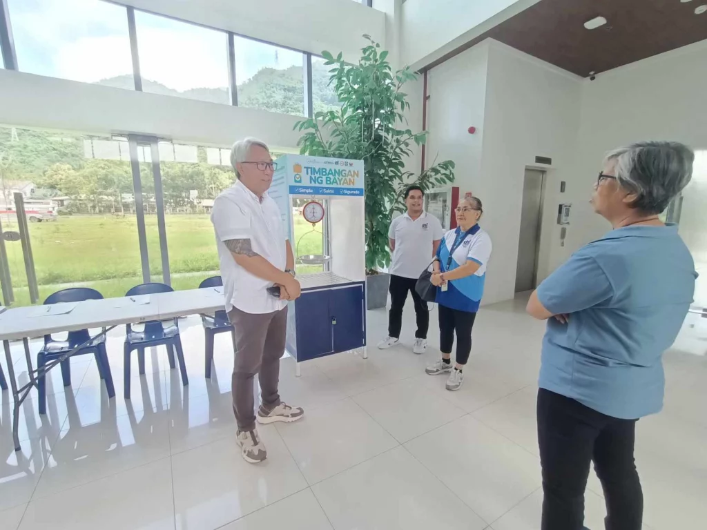 A group of officials from the Department of Trade and Industry (DTI) and Rizal Commercial Banking Corporation (RCBC) stand near a weighing scale setup labeled "Timbangan ng Bayan" during the launch event in Pakil, Laguna. The event is held in a bright, modern building with large windows and a view of greenery outside. The officials are engaged in conversation, and a table with chairs is visible in the background.