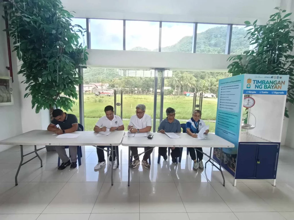 A group of officials from the Department of Trade and Industry (DTI) and Rizal Commercial Banking Corporation (RCBC) are seated at a table during the launch of the "Timbangan ng Bayan" in Pakil, Laguna. The event, held at the New Government Center, Tavera, features a ceremonial signing of acknowledgement and undertaking. A large poster with the "Timbangan ng Bayan" logo and information is displayed on the right side of the image. The background shows a scenic view of greenery and mountains through large windows.