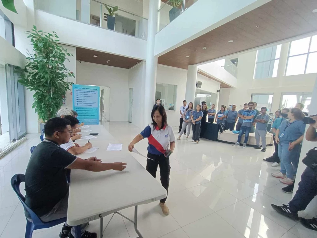 The image shows a launch event for the "Timbangan ng Bayan" initiative by the Department of Trade and Industry (DTI) and Rizal Commercial Banking Corporation (RCBC) in Pakil, Laguna. The event is held in a spacious, modern building with high ceilings and large windows. Several people are seated at a long table, signing documents, while a person stands in front of the table. In the background, a group of attendees, including officials and guests, stand and observe the proceedings. A large plant and a blue informational poster are visible on the left side of the image.