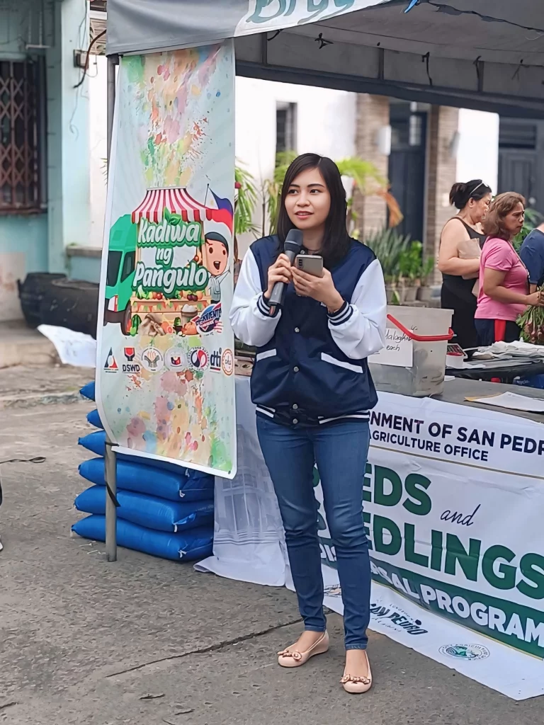 This image shows a person speaking at an event organized by the Department of San Pedro Agriculture Office, specifically for the "Seeds and Seedlings Distribution Program." The event appears to be part of the "Kadiwa ng Pangulo" initiative, which is indicated by the banner. This image is relevant as it depicts a community event focused on agricultural support and distribution of resources.