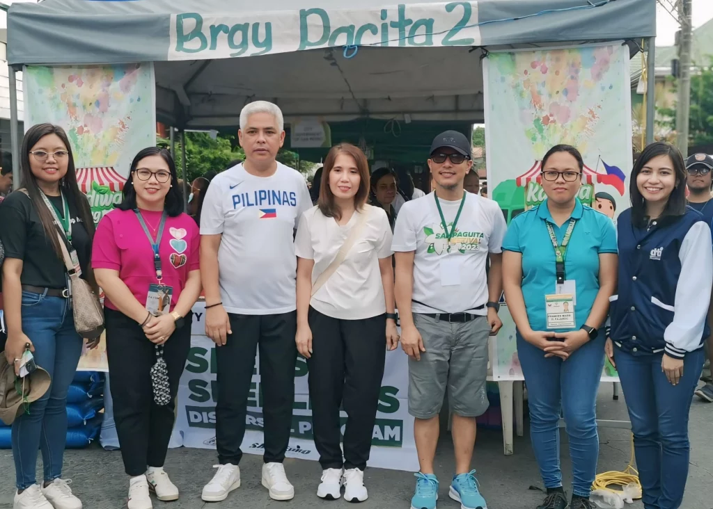 A group of seven individuals standing in front of a booth with a banner that reads "Brgy Pacita 2" at a community event. The individuals are dressed in various outfits, including shirts with logos and text such as "PILIPINAS" and "Sampaguita Festival 2023." The booth and banners in the background suggest participation in a local initiative, likely related to the Kadiwa ng Pangulo program, promoting consumer access to high-quality produce and manufactured products.
