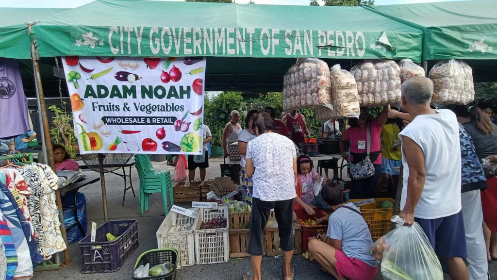 A market stall under a green tent labeled "CITY GOVERNMENT OF SAN PEDRO" displays a sign reading "ADAM NOAH Fruits & Vegetables WHOLESALE & RETAIL." The stall offers various fruits and vegetables, with several people browsing and purchasing items. Hanging bags of produce are visible, and there are crates filled with different types of fruits and vegetables on the ground. Clothing items are also displayed on the left side of the image. The scene is busy with multiple people interacting and shopping.