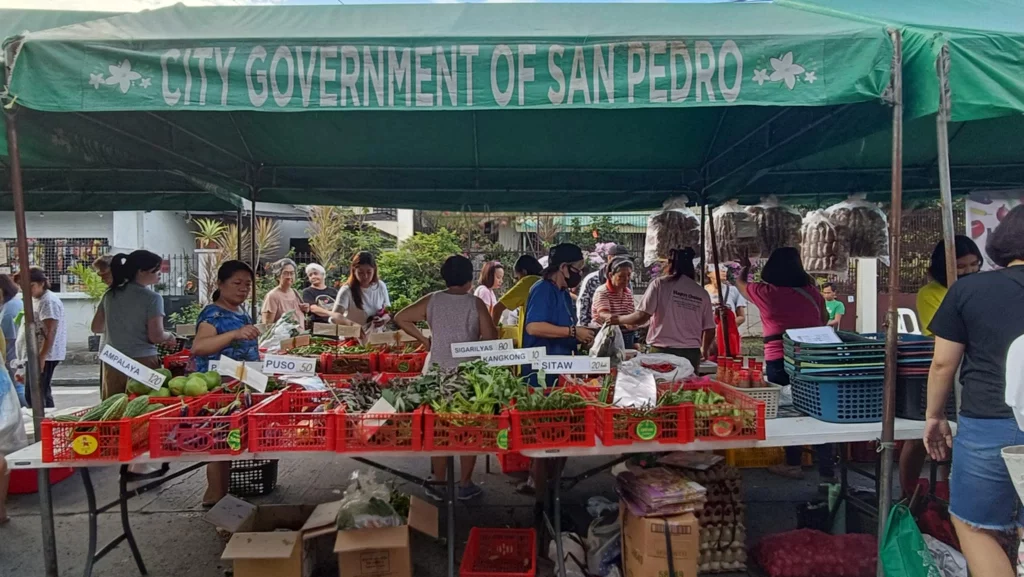 This image shows a market stall set up by the City Government of San Pedro, featuring a variety of fresh vegetables and produce. The scene is bustling with activity as people shop and interact with the vendors.