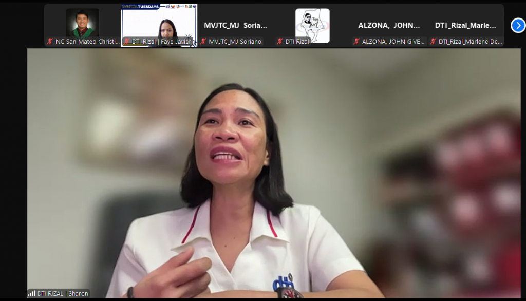A medium shot of a woman with short dark hair and a white collared shirt with a "DTI" logo, speaking during a Zoom meeting. The Zoom interface shows other participants' names and profile pictures at the top.