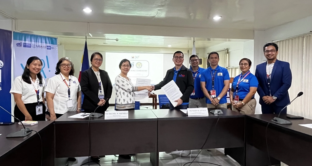 DTI Region 6 Regional Director Rachel N. Nufable and DepEd Region 6 Regional Director Ramir B. Uytico stand in the middle of a group, shaking hands and holding a signed memorandum of agreement. They are surrounded by representatives from both DTI and DepEd at a conference table, with the DTI logo in the background, during an event at the DTI Regional Office 6 on March 26, 2025.