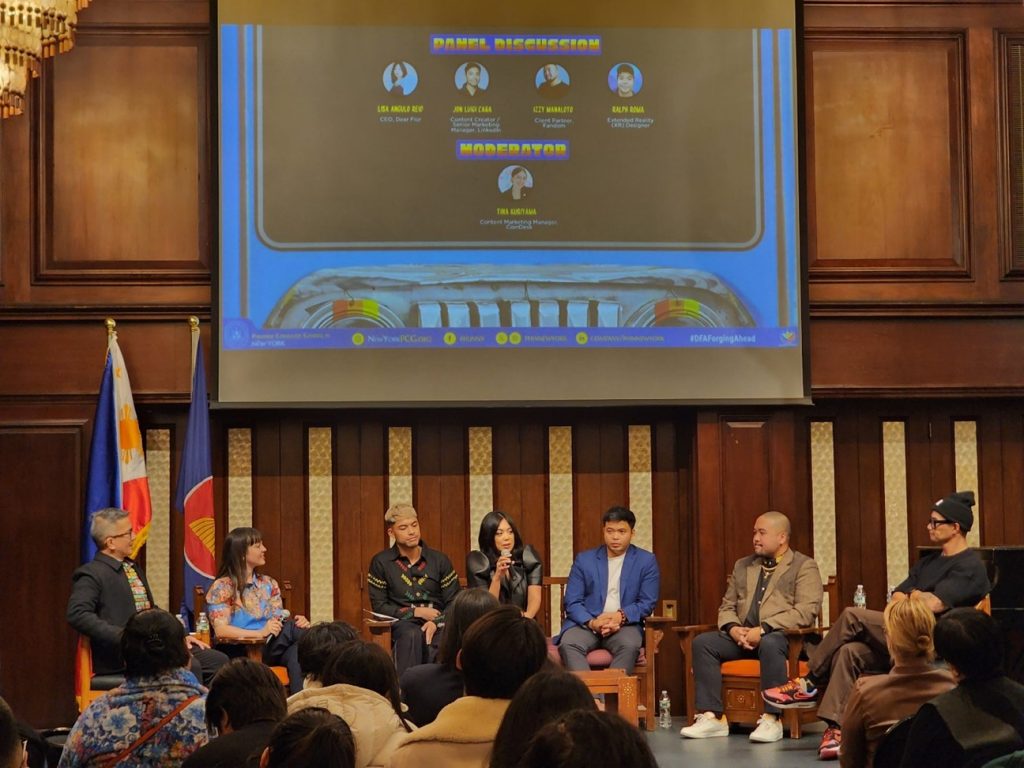 A group of Filipino and Filipino American professionals sits on a stage in front of a wooden backdrop, participating in a panel discussion. A large screen behind them displays the event title “AD-vancing Filipinnovation: Filipinos as a Global Creative Force.” Two Philippine flags are visible on the left, and audience members are seated in the foreground.