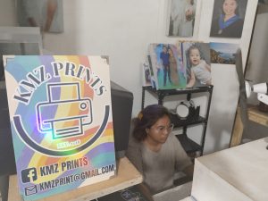 A printing shop display featuring a sign that reads “KMZ PRINTS” in a colorful design, with contact details. A woman wearing glasses sits behind a desk, surrounded by sample prints and portrait photographs on the walls.