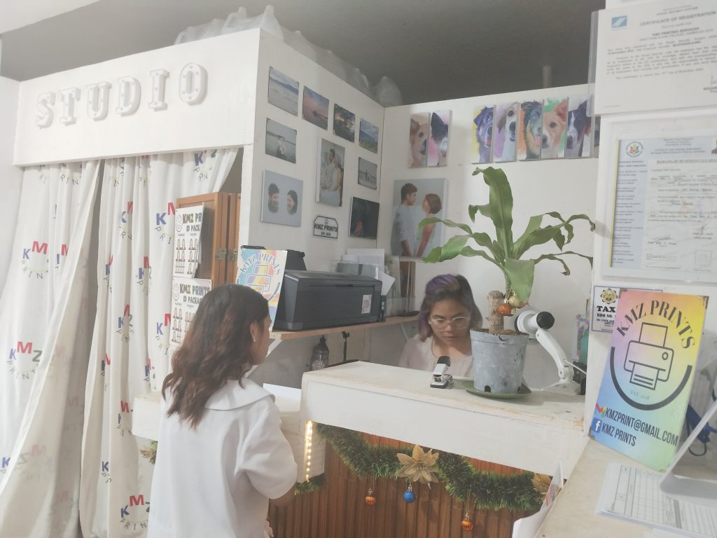 Interior of a photo printing studio with a “STUDIO” sign overhead. A woman with purple-tinted hair is behind the counter working on a computer, while a customer stands in front of the desk. Portrait samples and decorations are displayed on the walls and counter, and a potted plant sits on the reception area.