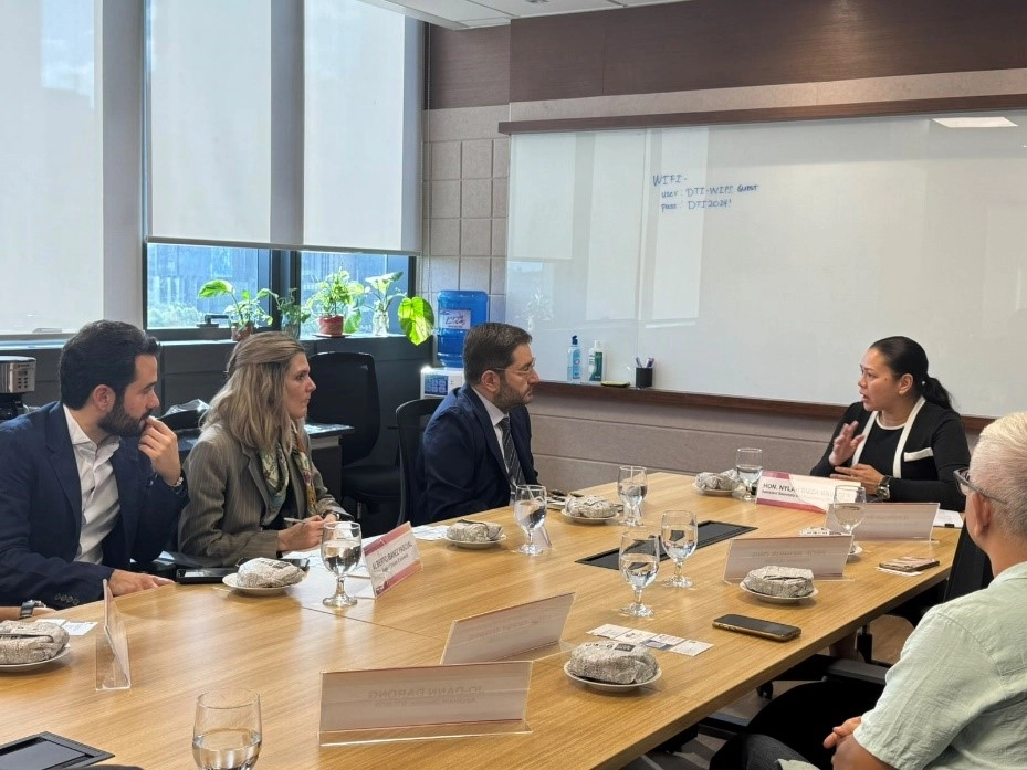DTI-CIG Assistant Secretary Nylah Rizza Bautista (right) sits at a conference table speaking with three representatives from IE University. They are in a meeting room with water glasses, nameplates, and a whiteboard in the background.