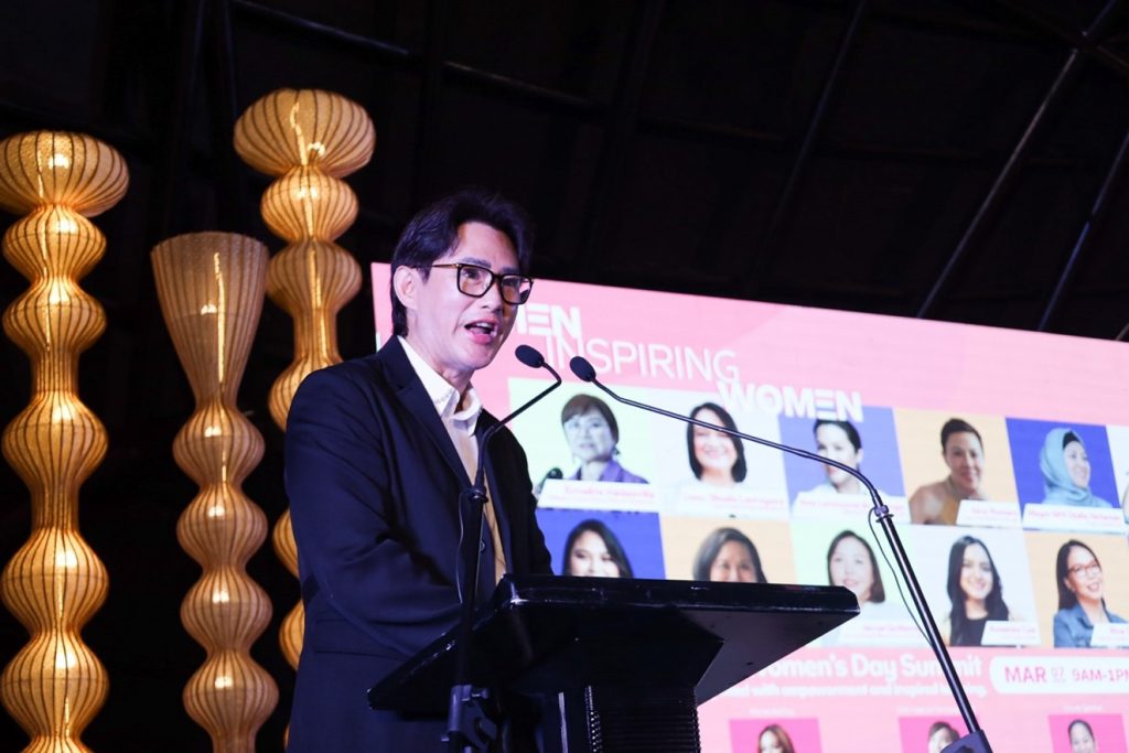 SM Supermalls President Steven Tan delivering a speech during the International Women's Day Summit at the Samsung Hall, SM Aura. Behind him is a lit presentation entitled, "Women Inspiring Women".