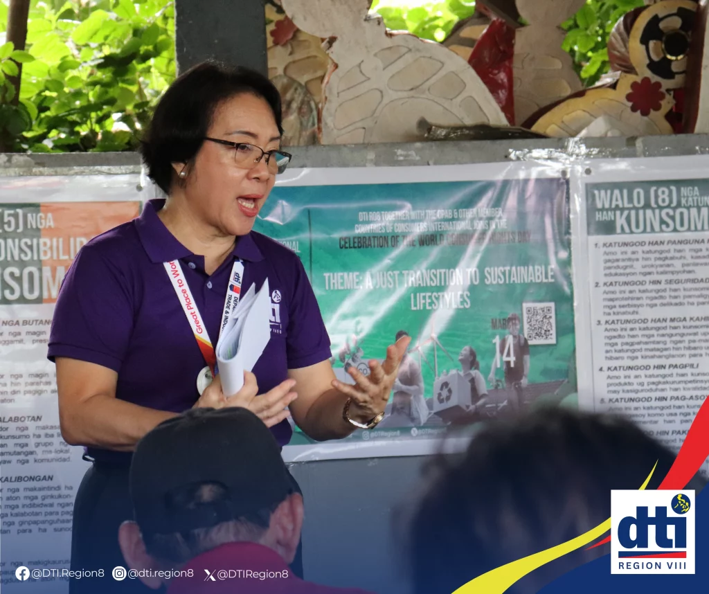 A DTI Region 8 representative wearing a purple shirt speaks to participants during an orientation on basic consumer rights and responsibilities in Barangay 79 Sirin, Tacloban City. Behind her, posters highlight the theme “A Just Transition to Sustainable Lifestyles.”