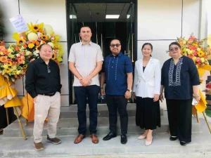 A group photo in front of a building with flower arrangements on each side, featuring (from left to right) an unidentified individual, Omega Global Technologies, Inc. President Zedric Ochoa (second from left), Omega GTI Asia, Inc. Executive Vice President Jayson Alfonso (center), DTI-Foreign Trade Service Corps Assistant Secretary Alma Argayoso (second from right), and DTI Region IV-A Regional Director Marissa Argente (rightmost).