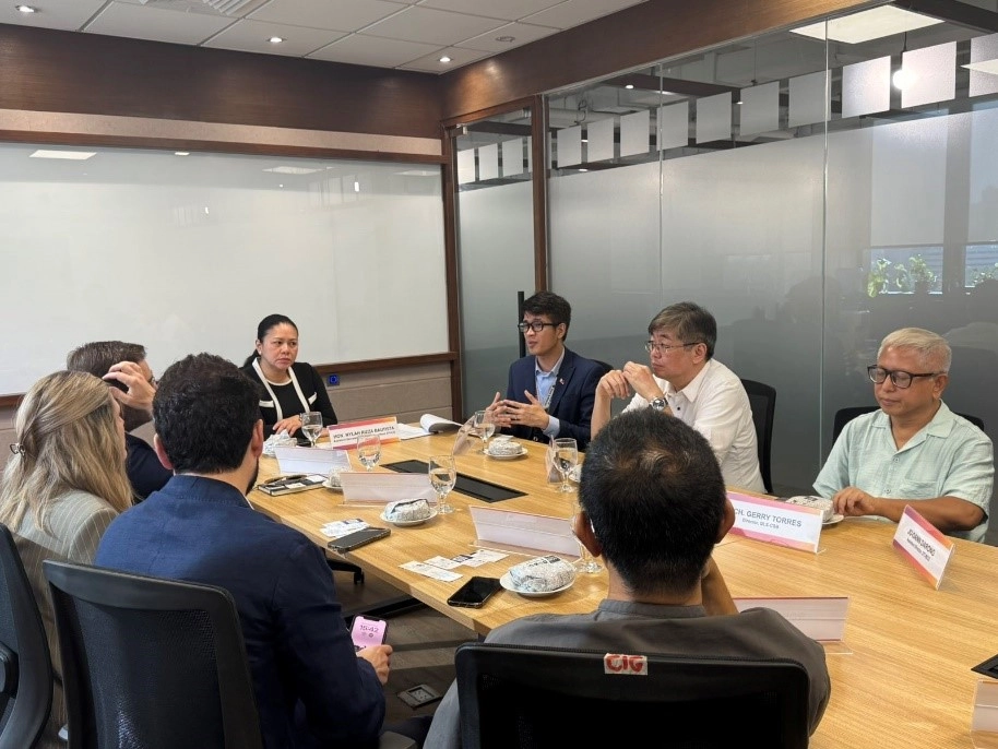 A group of representatives from DTI, IE University, and De La Salle-College of Saint Benilde sits around a conference table discussing creative industry collaborations. Nameplates and water glasses are on the table, and a glass partition and whiteboard are visible in the background.