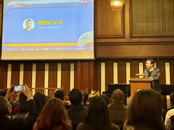 A man stands behind a wooden podium in a wood-paneled event hall, delivering closing remarks. A screen behind him displays his name, “Benedict M. Uy,” and the title “Trade Commissioner,” while audience members watch and take photos in the foreground.