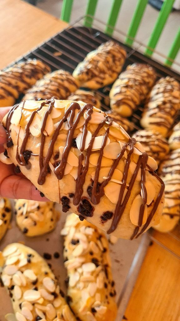 Sweet bread roll with chocolate drizzle and almond slices, held in hand, with other rolls on a cooling rack.