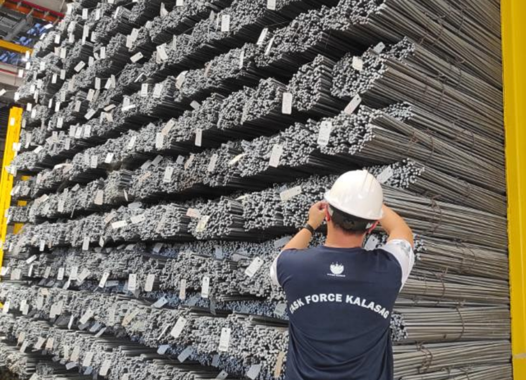 Steel rebar stacks in warehouse; inspector adjusts hardhat. DTI's E-KALASAG operation finds non-compliant products.