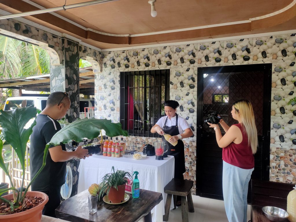 Rosemarie Guzman demonstrating sukang tuba prep; two videographers film the action on a porch.