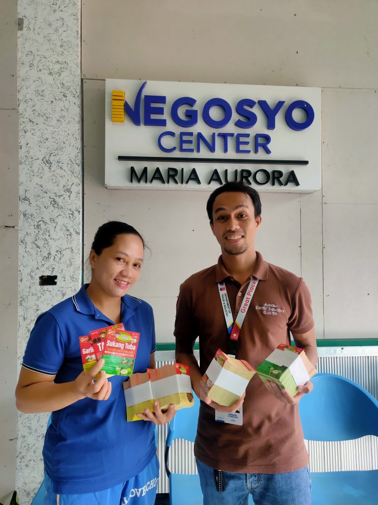 Two people stand in front of the "Negosyo Center Maria Aurora" sign, holding food products and boxes for business promotion.
