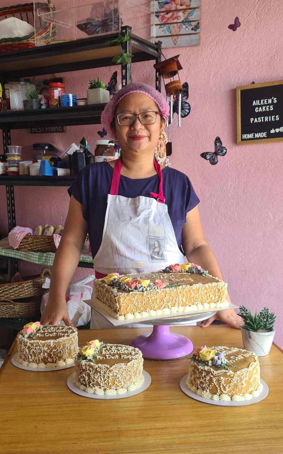Aileen, a baker, smiling behind a selection of iced caramel cakes in front of a pink wall decorated with butterflies.