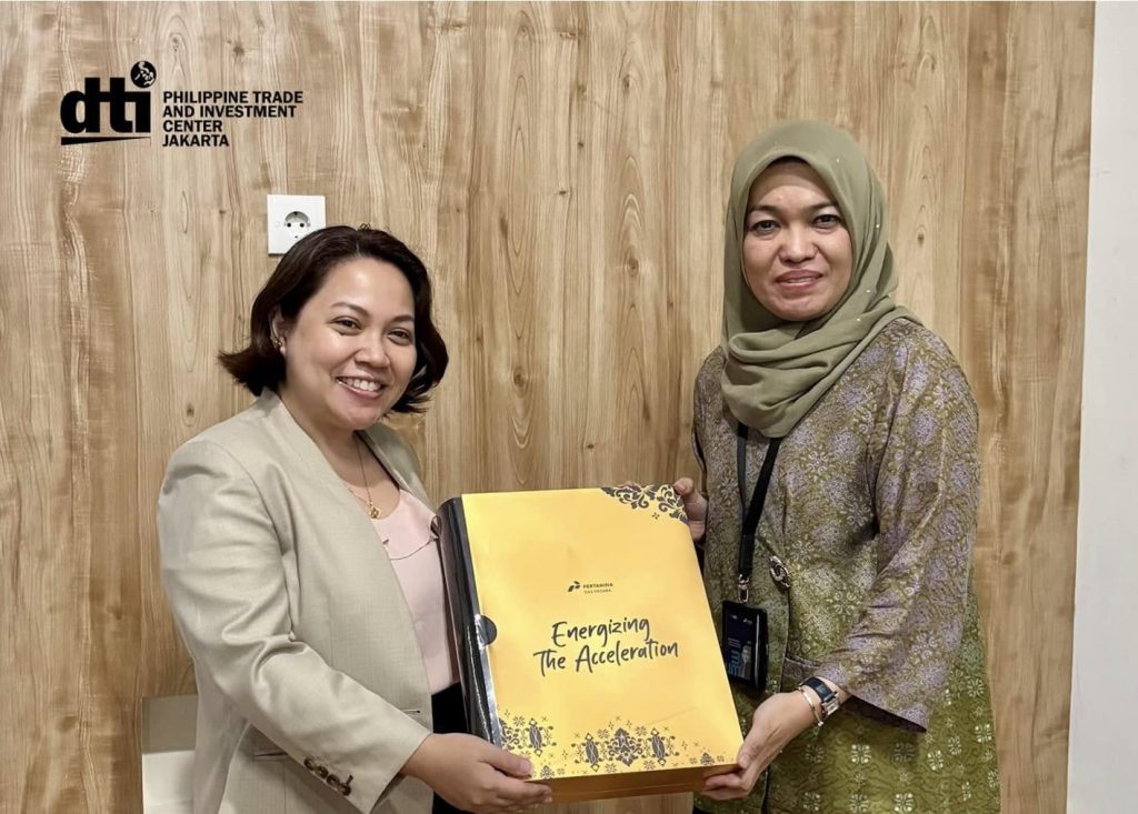 Two women stand side by side against a wooden wall, both smiling. One is holding a large folder with the text “Energizing the Acceleration.” They are identified as Philippine Trade and Investment Center (PTIC) in Jakarta’s Commercial Counsellor, Jollan Margaret Llaneza (left), and Pertamina Gas Negara’s Director of Strategy and Business Development, Rosa Permata Sari (right), meeting on January 23, 2025.