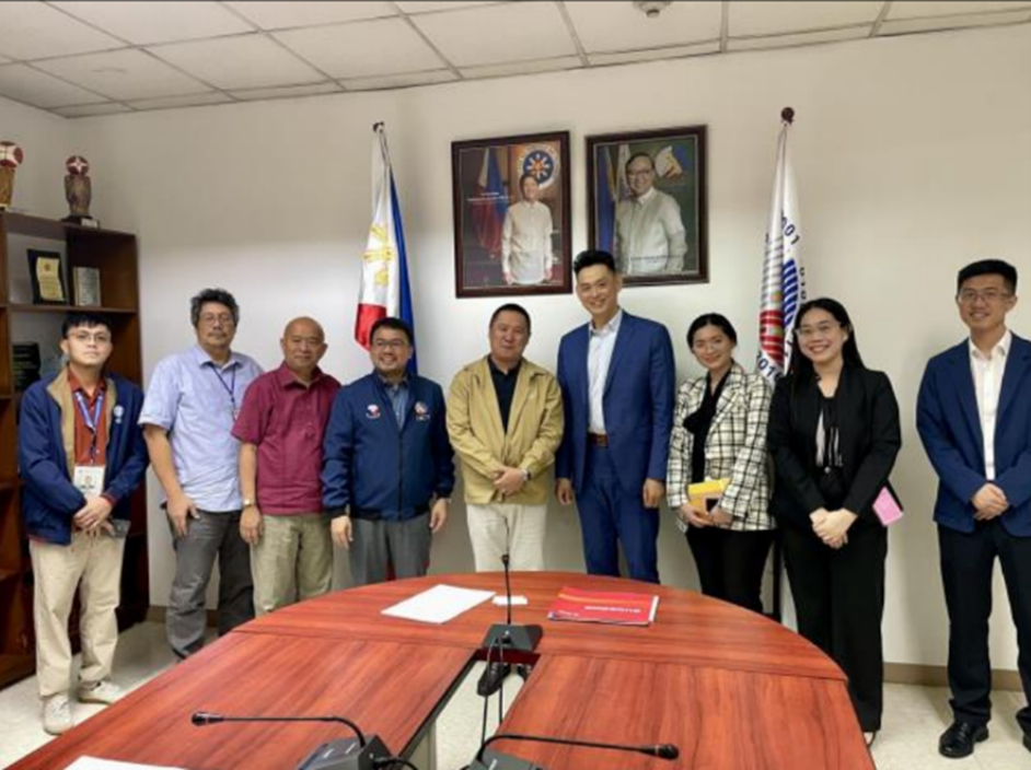 Group photo taken in a conference room, with DICT-OUIMCU Undersecretary Jeffrey Ian Dy in the center, alongside other DICT representatives; Guodong Group CEO Longke Hu (fourth from right); DTI-FTSC Regional Coordinator for Europe and Middle East Rain Anne Mojica (third from right); DTI-FTSC Regional Coordinator for Chinas and East Asia Melody Dicen (second from right); and Guodong Group Sales Representative Ning Li (leftmost), in front of Philippine flags and official portraits.