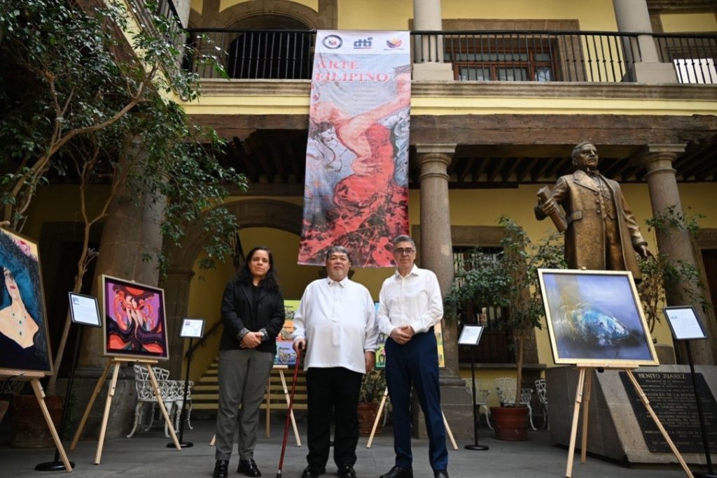 Three representatives from the Philippine Trade and Investment Center in Mexico (from left to right: Trade Assistant Liliana Calderon, Commercial Counsellor John Paul Iñigo, and Trade Assistant Luis Castro) stand in a historic courtyard. Several colorful paintings are displayed on easels around them, and a large statue is visible on the right. A banner above reads 'ARTE FILIPINO.
