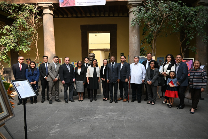 A group of around 15 people, including officials from the Philippine Trade and Investment Center in Mexico, pose in a historic courtyard with tall columns and greenery. A statue stands behind them, and colorful artwork is visible on display in the background.
