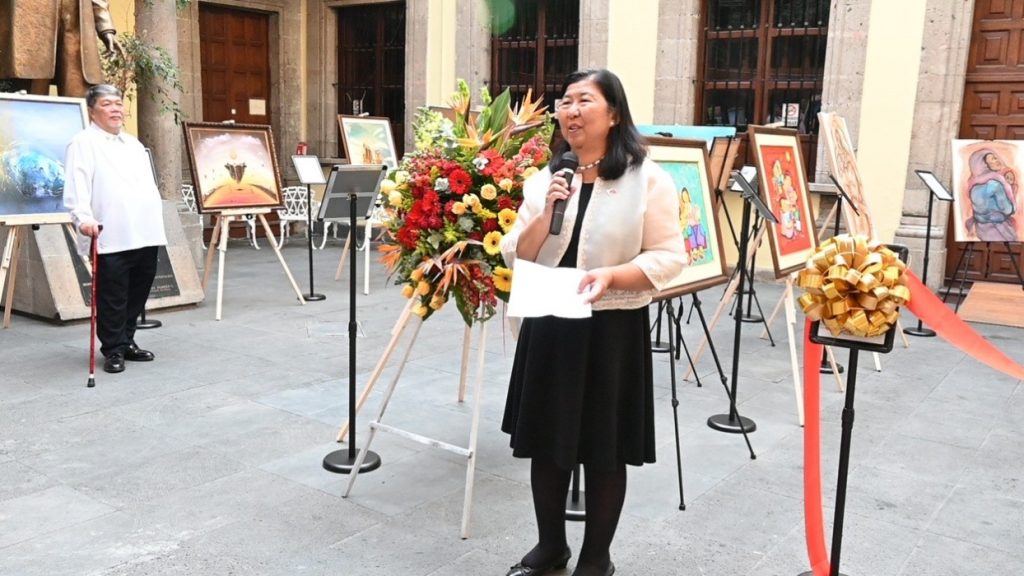 Her Excellency Lilybeth R. Deapera, Philippine Ambassador to Mexico, stands holding a microphone and giving opening remarks in a courtyard. Colorful paintings are displayed on easels behind her, alongside a floral arrangement and a ribbon. A man in white attire looks on from the background.