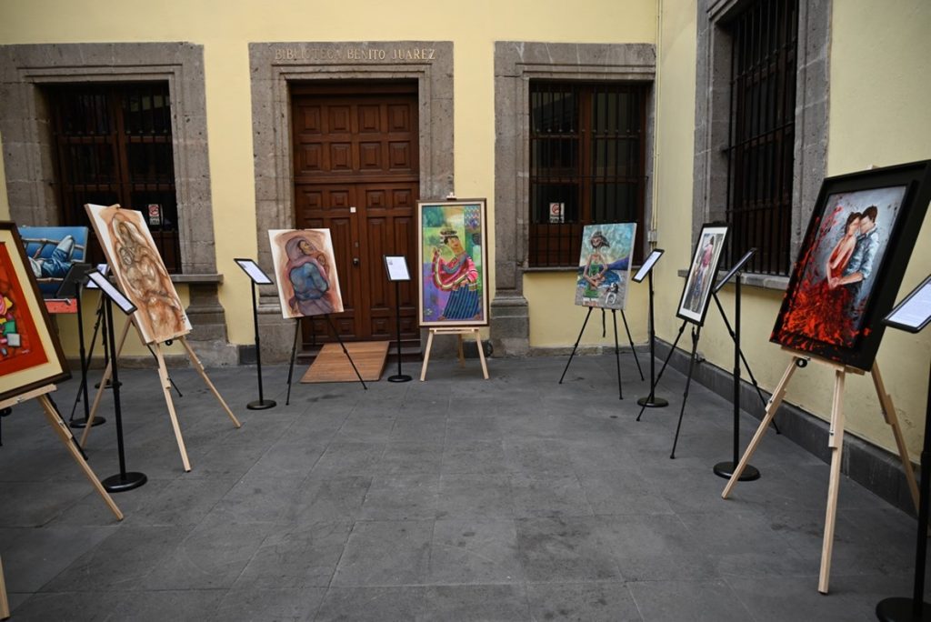 Several vibrant paintings displayed on easels in a courtyard, arranged in front of a wooden doorway. Each artwork showcases a unique style and subject, representing part of the 30-piece Filipino Art Nook exhibition.