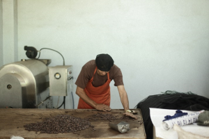 Man in orange apron sorts cocoa beans on a wooden table next to chocolate processing equipment.