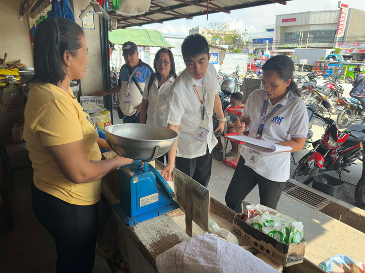 At a busy market in SOCCSKSARGEN, Philippines, two officials from the Department of Trade and Industry (DTI) monitor rice prices. One official in a white uniform shirt and ID badge holds a clipboard looking at it, while the other official in a white shirt stands nearby, observing a vendor in a yellow shirt. who is using an analog scale to weigh rice.