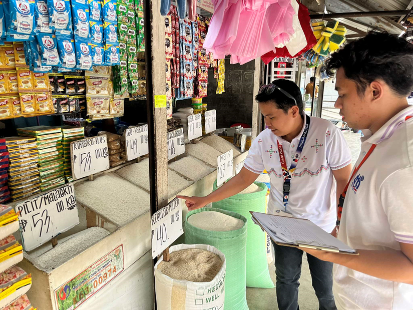 Two Department of Trade and Industry (DTI) officers conduct price monitoring in a public market for imported rice. One officer points at a sign displaying the price while the other takes notes on a clipboard. The market stall offers various rice types and other goods such as instant noodles and drinks.