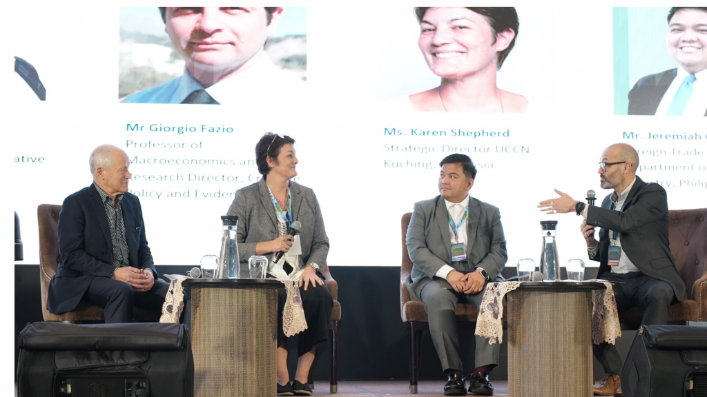 Four panelists are seated on a stage in armchairs, facing the audience. A large screen behind them shows their names, photos, and titles (including “Mr Giorgio Fazio,” “Ms Karen Shepherd,” and others). The panelists are dressed in business attire, with the man on the far right holding a microphone and gesturing as he speaks. Small tables with water glasses sit in front of each panelist. They are engaged in a discussion about a regional approach to the creative economy.