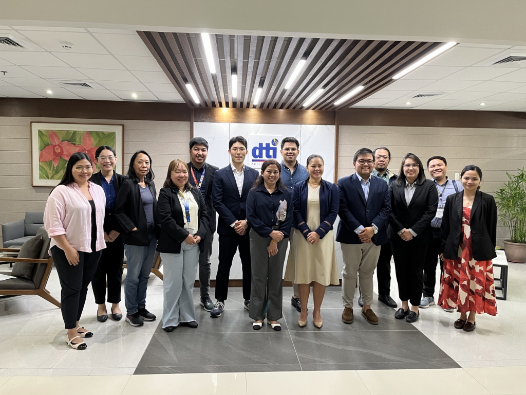 A group of twelve individuals stands together in a well-lit office lobby, in front of a wall bearing the DTI logo. They are dressed in business attire and smiling at the camera. The group includes representatives from the Department of Trade and Industry, Competitiveness and Innovation Group, Export Marketing Bureau, Board of Investments, Department of Environment and Natural Resources, and the ASEAN-Korea Centre.